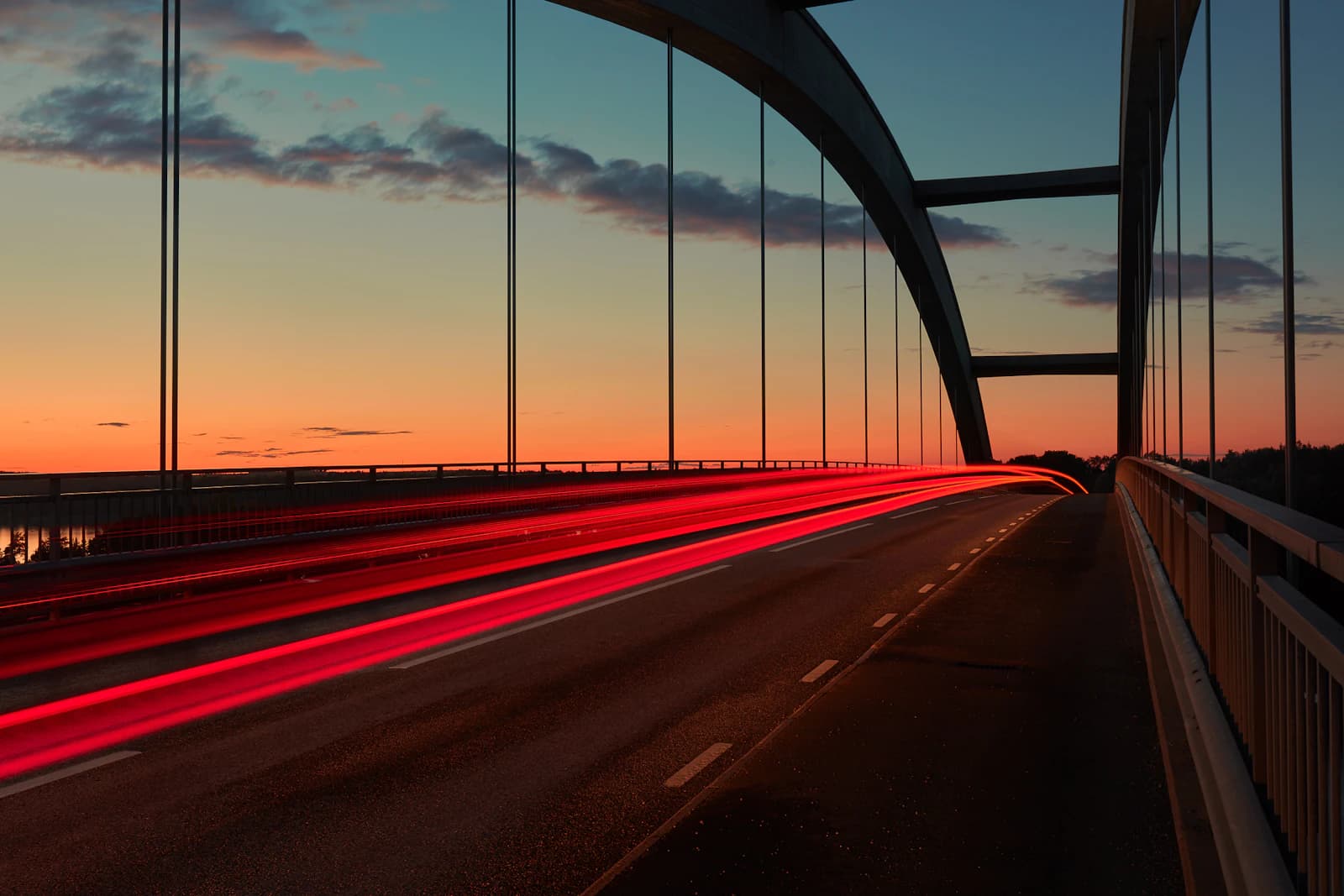 Long-exposure photo of red car light trails along a road on an arched bridge at sunset