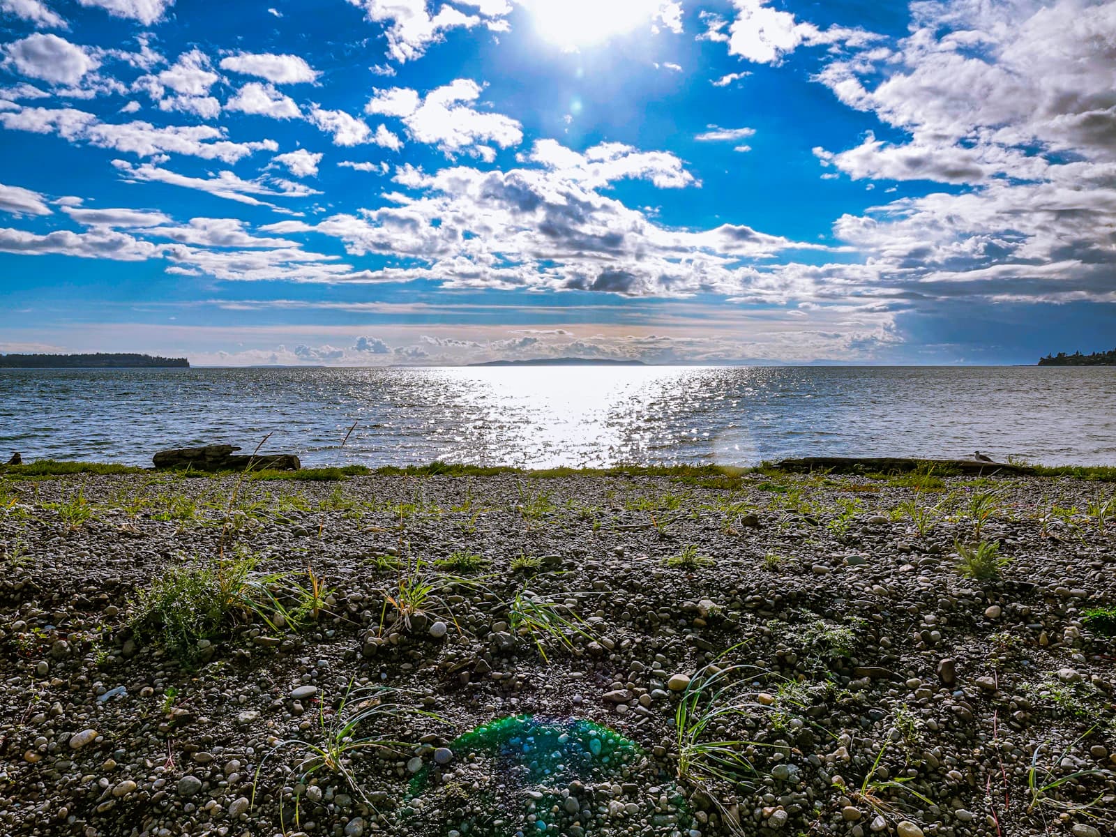 Blue sky with clouds over a pebble lakeshore and sunlight sparkling on the water