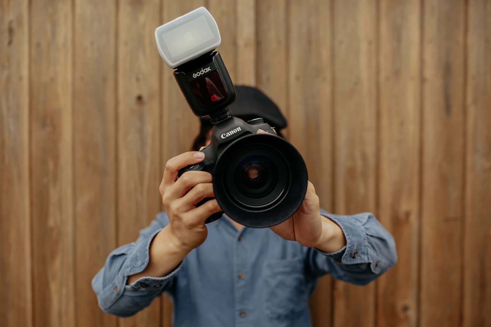 Person hiding behind a flash-equipped camera in front of a wooden wall